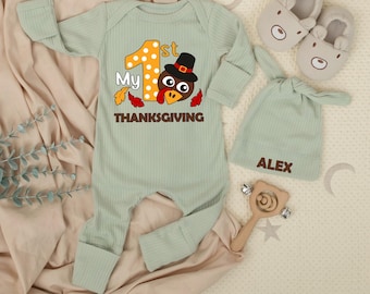 May include: A light green baby outfit set with a Thanksgiving theme. The set includes a long-sleeved onesie with a turkey graphic and the text "My 1st Thanksgiving", a matching hat with the name "ALEX", and bear-shaped slippers.