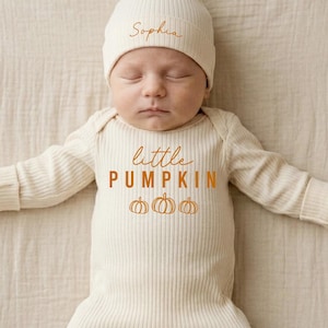 May include: A cream-colored ribbed long-sleeve baby sleeper and matching beanie. The sleeper features the text "little PUMPKIN" in orange, with three small orange pumpkin illustrations below. The beanie is personalized with the name "Sophia" in orange.