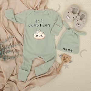 May include: A light green baby outfit set with a long-sleeved onesie, a knotted hat, and bear-shaped slippers. The onesie has the text "lil dumpling" and a dumpling graphic. A wooden rattle is also included.