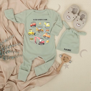 May include: Sage green baby onesie with construction vehicle graphics and the text "GOD SAYS I AM". Includes a matching hat, bear-shaped slippers, and a wooden rattle. The onesie has long sleeves and ribbed texture.
