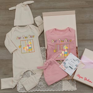 May include: A baby gift set featuring a cream-colored and pink ribbed onesie and hat, with the text "Got Too Silly" and a duck graphic. The set includes a gift box and a welcome card.
