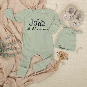 May include: A light green baby outfit set, including a ribbed jumpsuit with "John William" printed on the front, a matching knotted hat, and a pair of bear-shaped slippers. A wooden rattle is also included.