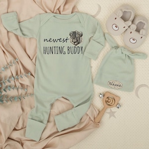 May include: A light green baby outfit set with the text "newest HUNTING BUDDY" and a deer graphic. The set includes a long-sleeved onesie, a hat, and a pair of bear-shaped booties. A wooden rattle is also included.