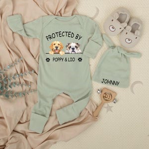 May include: A light green baby outfit set with a long-sleeved onesie, a knotted hat, and bear-shaped slippers. The onesie features the text "PROTECTED BY" above illustrations of a golden retriever and a bulldog, with the names "POPPY & LIO" below.