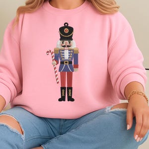 Könnte beinhalten: Ein hellrosa Sweatshirt mit einem gestickten Nussknacker-Soldaten-Design. Der Nussknacker trägt eine blaue Uniform mit goldenen Akzenten, eine rote Hose und schwarze Stiefel und hält eine Zuckerstange. Das Design ist mittig auf dem Sweatshirt platziert.