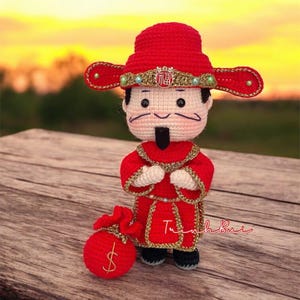May include: A handmade crochet doll of a Chinese God of Wealth, dressed in a red robe with gold trim and a matching hat. The doll holds a red money bag with a gold dollar sign. The doll is standing on a wooden surface.