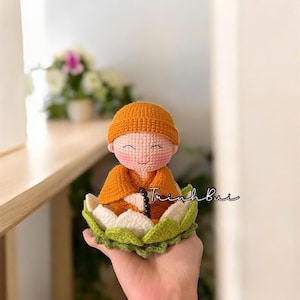 Può includere: Bambola all'uncinetto fatta a mano di una figura meditante, che indossa una veste e un cappello arancioni. La bambola è seduta su un fiore di loto bianco e verde. La bambola ha un viso sorridente e perline nere. Il testo "Trinh Buc" è scritto sul fiore di loto.
