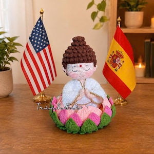 May include: A crocheted figurine of a person with brown hair in a bun, wearing a white robe with gold trim, sitting in a pink and green lotus flower. The figure has a red dot on the forehead and hands in a prayer position. The text TrinhBui is visible.