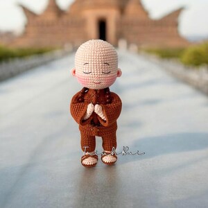 May include: A handmade crochet doll of a Buddhist monk in a brown robe, with hands clasped in prayer. The doll has a bald head, rosy cheeks, and closed eyes. The background features a blurred temple and the artist's signature.