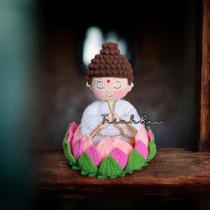 Pode incluir: Figura de Buda de crochê com cabeça castanha, túnica branca e um ponto vermelho na testa. O Buda está sentado em uma flor de lótus com pétalas rosa, verdes e brancas. A figura tem um colar de corrente dourada.