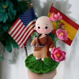 Puede incluir: Muñeca de monje de ganchillo con túnica marrón y collar de cuentas, con una expresión serena. Se encuentra sobre una base de hoja de nenúfar verde, con flores de loto rosas y pequeñas banderas de Estados Unidos y España al fondo.