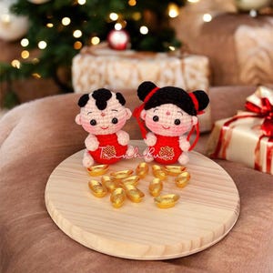 Puede incluir: Muñecos de ganchillo de un niño y una niña con trajes rojos, pelo negro y mejillas rosadas, sentados sobre una tabla de madera. Lingotes de oro los rodean. Las muñecas son probablemente hechas a mano y son una decoración festiva.