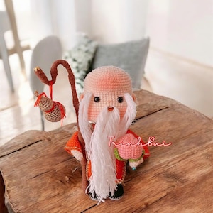 May include: A handmade crochet doll of a wise old man with a long white beard, holding a staff and a pink pouch. The doll is wearing an orange and red robe. The text "Trinh Bui" is written on the wooden surface.