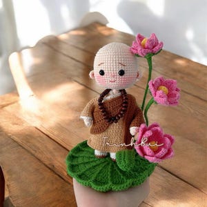 Op de afbeelding: Een gehaakte monnikfiguur in een bruine robe en kralenketting, staand op een groen lelieblad. Roze lotusbloemen met gele centra bevinden zich rechts. De figuur heeft een lachend gezicht en houdt het lelieblad vast.