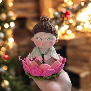 May include: A handcrafted crochet doll of a meditating figure, seated in a pink lotus flower. The doll has a brown hair, a white shawl, and a serene expression. The lotus flower has pink petals and green leaves. The doll is approximately 4 inches tall.