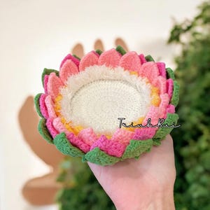 Puede incluir: Un posavasos de ganchillo hecho a mano con forma de flor. El posavasos presenta hilo rosa, verde, amarillo y blanco. El posavasos se sostiene en una mano. El texto "Trinh Bui" está escrito en el posavasos.