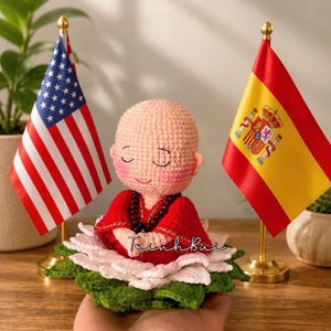 May include: A handmade crochet doll depicting a meditating figure in a red robe, seated on a white and green lotus flower. The doll has closed eyes and a peaceful expression. The American and Spanish flags are in the background.