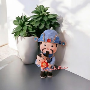 Può includere: Bambola all'uncinetto fatta a mano di un uomo con cappello e abito blu, che tiene in mano una bambola più piccola. La bambola ha baffi e barba neri, con dettagli dorati e rossi. Lo sfondo include un vaso bianco con piante verdi.