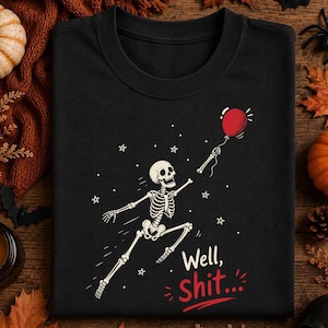 May include: A black crew neck t-shirt featuring a graphic of a white skeleton running while holding a red balloon. The design includes small white stars and the text "Well, Shit..." printed in white and red lettering below the skeleton figure.