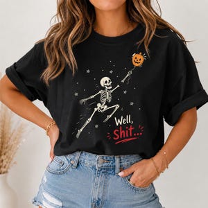 May include: A black short-sleeved t-shirt featuring a white graphic of a skeleton reaching for an orange pumpkin balloon. The text "Well, Shit..." is printed in white and red below the skeleton, surrounded by small white star accents.