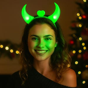 Può includere: Una donna indossa una fascia verde luminosa con corna curve e un dettaglio a forma di cuore. La fascia emette una luce verde, che si riflette sul viso della donna. Lo sfondo presenta luci bokeh soffuse.