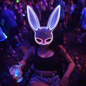 May include: A white bunny mask with glowing neon pink and blue outlines. The mask is worn by a person in a black tank top and denim shorts, seated in a chair. The background is a crowded, dimly lit event.