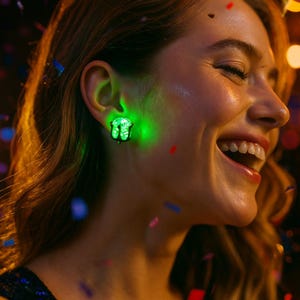 May include: A woman wearing a glowing green earring. The earring is a small, ornate design that emits a bright green light. The woman is smiling, and confetti is falling around her. The background is blurred with warm, golden tones.