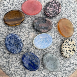 Könnte beinhalten: Eine Sammlung polierter Edelsteine in verschiedenen Farben und Mustern. Die Steine umfassen Tigerauge, Rosenquarz, Rhodonit, Lapislazuli und Dalmatinerjaspis. Die Steine sind oval geformt und werden auf einer grau strukturierten Oberfläche präsentiert.