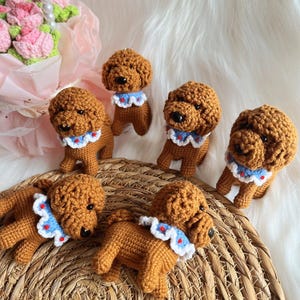 May include: A collection of handmade crochet dog figurines. The dogs are brown with black eyes and noses, wearing blue and white ruffled collars. They are arranged on a woven surface with a bouquet of pink flowers in the background.
