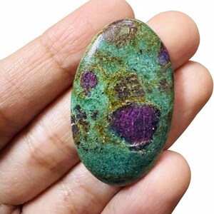 Op de afbeelding: Een ovale edelsteen met een turquoise basiskleur, bespikkeld met donkerbruine en levendige paarse insluitsels. De gepolijste steen wordt in een hand gehouden, wat de natuurlijke schoonheid en unieke kleurvariaties benadrukt. Ongeveer 5 cm lang.