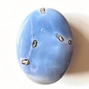 May include: An oval-shaped, polished blue gemstone with a smooth, glossy surface. The stone features several small, dark, irregular inclusions scattered across its light blue, semi-translucent body, creating a distinct contrast in texture.
