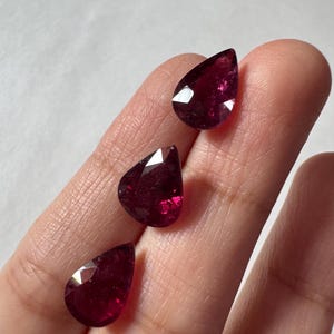 May include: Three loose, pear-shaped faceted gemstones with a deep, translucent magenta-red color. Each stone features a pointed top and a rounded base, displaying multiple geometric facets that reflect light across their polished surfaces.