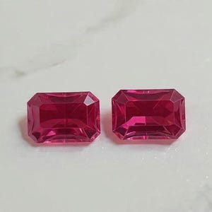 Op de afbeelding: Twee rechthoekige, gefacetteerde, dieproze edelstenen. De edelstenen liggen op een wit marmeren oppervlak. De edelstenen hebben een levendige, verzadigde kleur en een hoge mate van helderheid.