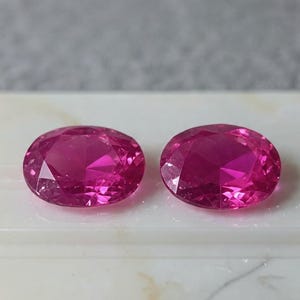 Può includere: Due gemme rosa vibranti, taglio ovale. Le gemme sono esposte su una superficie bianca, con uno sfondo morbido e sfocato. Le sfaccettature delle pietre catturano la luce, creando un effetto scintillante. Queste gemme sono probabilmente per la creazione di gioielli.