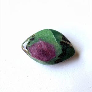 Può includere: Una gemma lucidata con un vibrante centro di rubino circondato da un esterno verde. La pietra ha una forma liscia e arrotondata e una finitura lucida. Il rubino è di un colore rosso intenso e ricco, mentre lo strato esterno è un mix di verde e nero.