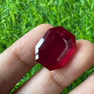 Könnte beinhalten: Ein tiefroter, facettierter Edelstein mit achteckiger Form. Der Stein besitzt eine polierte, durchscheinende Oberfläche mit scharfen, geometrischen Kanten, die das Licht reflektieren und einen satten, kräftigen Karmesinton zeigen.