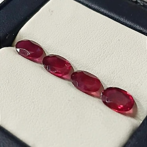 Peut inclure: Quatre pierres précieuses ovales, d'un rouge profond, sont présentées dans un écrin noir avec un intérieur blanc. Les gemmes sont alignées, mettant en valeur leurs surfaces polies et leur couleur riche. L'écrin offre un arrière-plan contrasté.