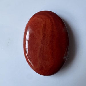 Può includere: Una pietra di diaspro rosso lucidata di forma ovale con una superficie liscia e brillante. La pietra presenta variazioni di colore terracotta e rosso mattone con sottili motivi vorticosi e una sottile linea orizzontale scura.