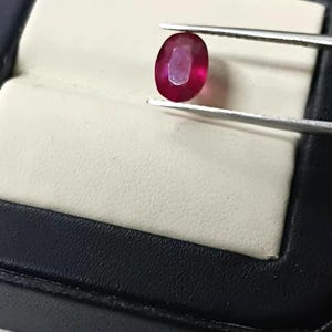 Könnte beinhalten: Ein lebendiger, oval geschliffener Rubin, der von einer Pinzette gehalten wird und vor einer schwarzen Schmuckschatulle mit cremefarbenem Innenraum präsentiert wird. Die tiefrote Farbe des Rubins ist deutlich sichtbar.