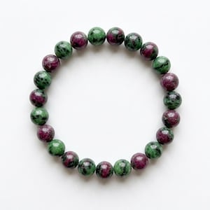 May include: A beaded bracelet featuring round gemstones in shades of green and maroon. The bracelet is made of polished stones, strung together to form a circular shape. The gemstones have a speckled appearance, with a mix of colors.