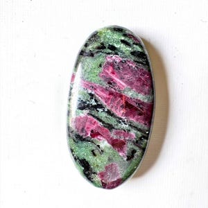 May include: An oval-shaped polished gemstone featuring a mottled pattern of deep magenta, forest green, and black mineral inclusions. The surface is smooth and reflective, displaying a natural crystalline texture throughout the stone's surface.