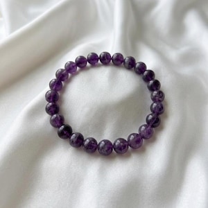May include: A bracelet made of polished, round amethyst beads. The deep purple beads are strung together, forming a complete circle. The bracelet sits on a white, silky fabric.