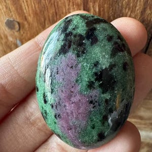 May include: An oval-shaped polished gemstone featuring a mottled surface with a mix of deep forest green, vibrant magenta, and dark black inclusions. The stone has a smooth, glossy finish that reflects light across its varied natural colour patterns.