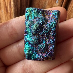 Puede incluir: Un espécimen mineral rectangular e iridiscente con una superficie rugosa y texturizada. La piedra muestra una vibrante gama de colores, incluyendo verde azulado, azul, púrpura y dorado. El mineral se sostiene en una mano, mostrando su belleza natural.