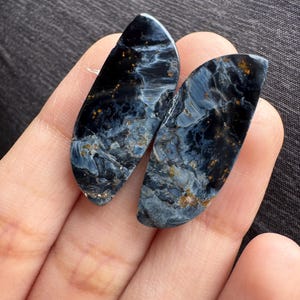 May include: Two polished pietersite gemstones. The stones are a mix of dark blue, light blue, and gold, with a swirling pattern. The gemstones are oval-shaped and are held in a hand.