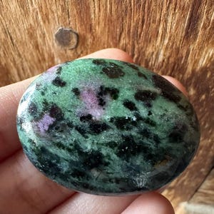May include: A polished oval-shaped zoisite stone featuring a mottled pattern of deep forest green, vibrant magenta-pink, and dark black inclusions. The surface is smooth and reflective, showcasing the natural mineral variations within the stone.