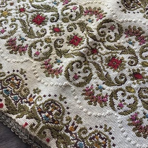May include: Close-up of a cream-colored textile with an intricate floral pattern. The design features red, pink, and blue flowers with green foliage. The edges are trimmed with a decorative border.