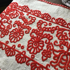 May include: Cream-colored textile with intricate red floral embroidery. The design features stylized flowers and scrolling vines, creating a decorative border. The edges are finished with a red zigzag stitch, adding a contrasting detail to the piece.