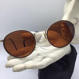 May include: A pair of round, brown-tinted sunglasses with a gold metal frame. The sunglasses are resting on a white hand.