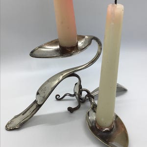 May include: A unique, handcrafted candle holder made from repurposed silverware. The silver-toned metal piece features two spoons holding white and peach-colored candles. The design includes curved stems and decorative flourishes, creating an elegant, vintage aesthetic.